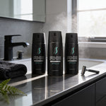 Three black deodorant cans with theAXE brand name and the word 'APOLLO' printed on them, placed on a bathroom counter with a razor and a black towel.
